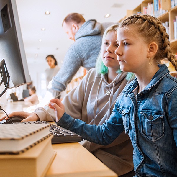 Een jong meisje in een spijkerjasje zit naast een andere persoon achter een computer in een bibliotheek. Op de voorgrond liggen boeken, en op de achtergrond zijn andere bezoekers en boekenplanken zichtbaar.